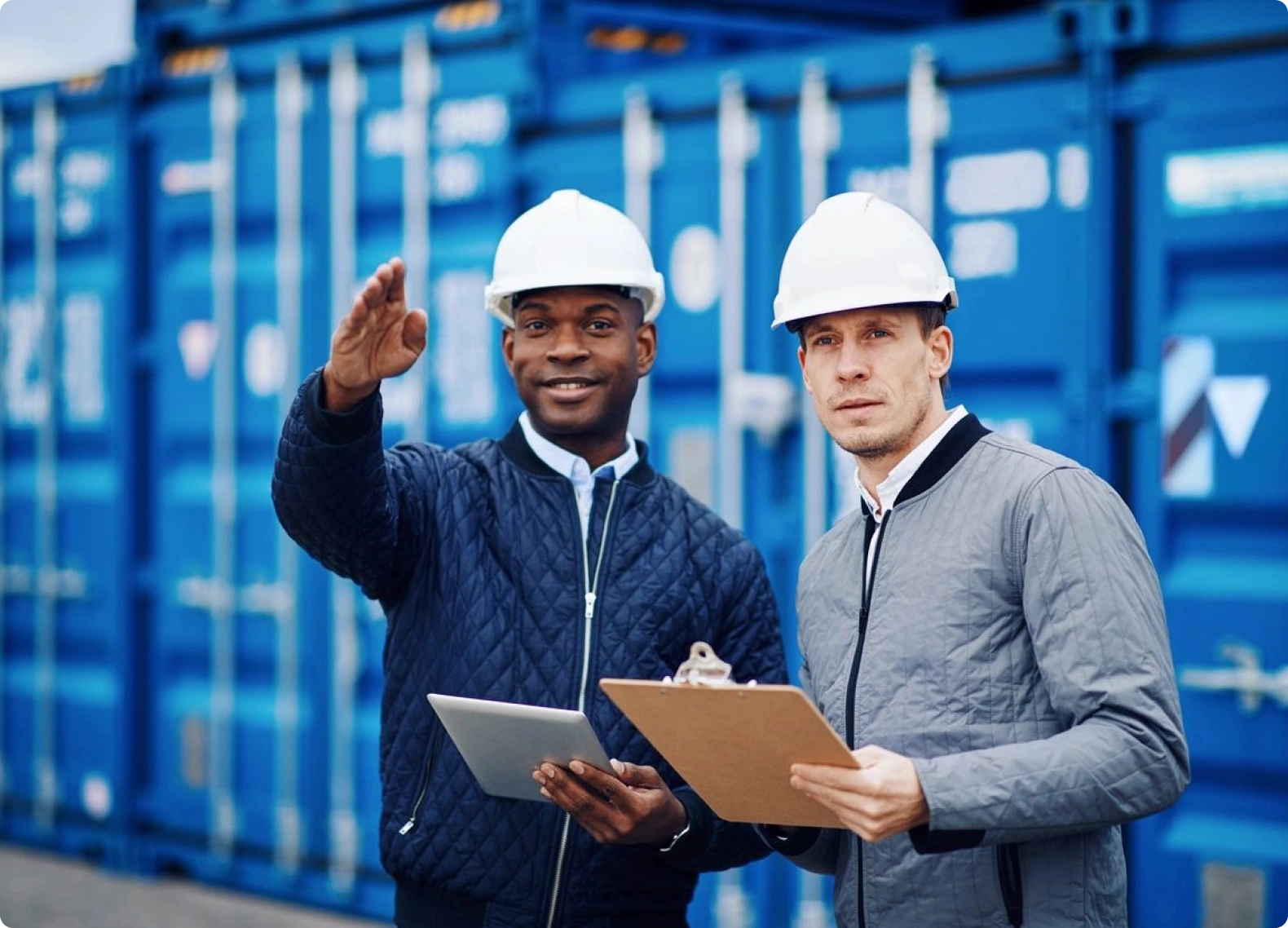 Two men in white hard hats holding clipboards.